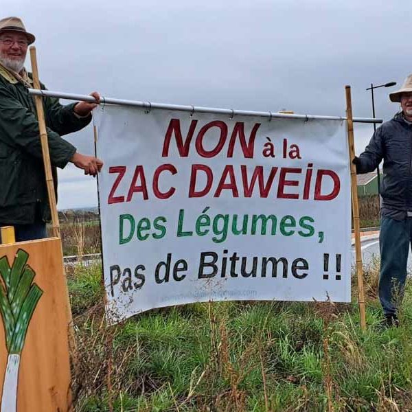 2 militants souriants sous un ciel pluvieux tiennent la banderole au milieu d'un rond-point près du Leclerc de Issenheim. Une pancarte poireau prend la pose avec eux.