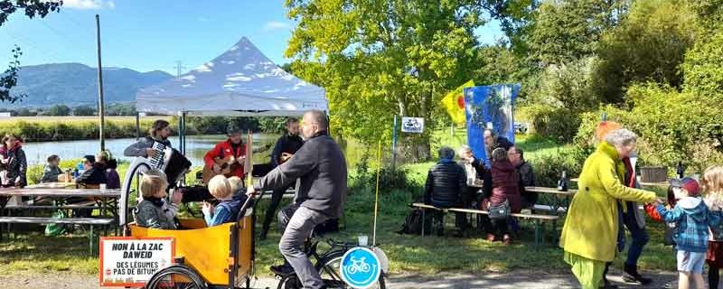 Vue sur des tables et bancs, le chapiteau et 3 musiciens, des personnes attablées et au premier plan un vélo-cargo qui transporte 4 enfants enthousiastes.