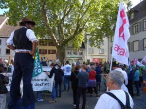 Place de la liberté, un homme monté sur échasses observe le discours, un autre tient un drapeau ATAC