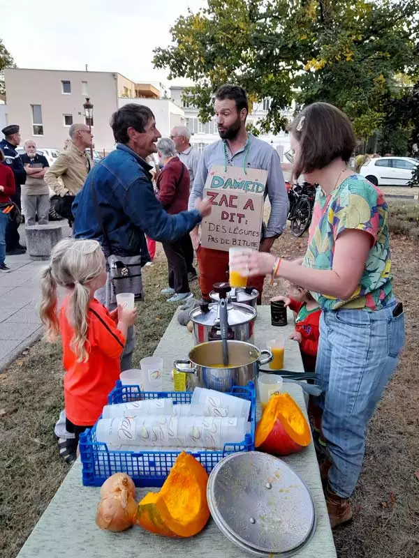 Distribution de soupe aux légumes d'automne. Au premier plan, 1 enfant et 3 adultes, et en arrière-plan un gendarme discute avec un membre du collectif.